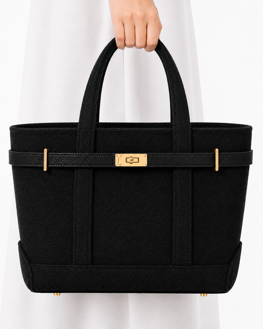 A black structured tote bag with gold-tone hardware, held by a person wearing a white dress against a clean white background.
