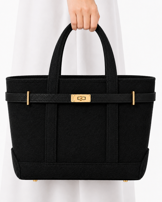 A black structured tote bag with gold-tone hardware, held by a person wearing a white dress against a clean white background.
