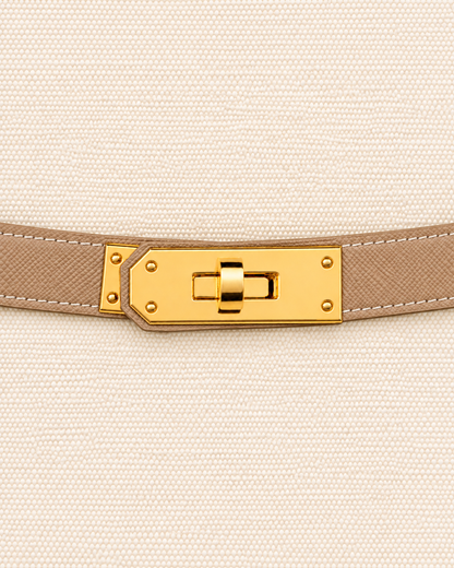 A close-up of a beige textured leather belt with a gold-tone turn-lock clasp, shown against a matching beige woven fabric background. Boat Tote Boatkin bag handbag 