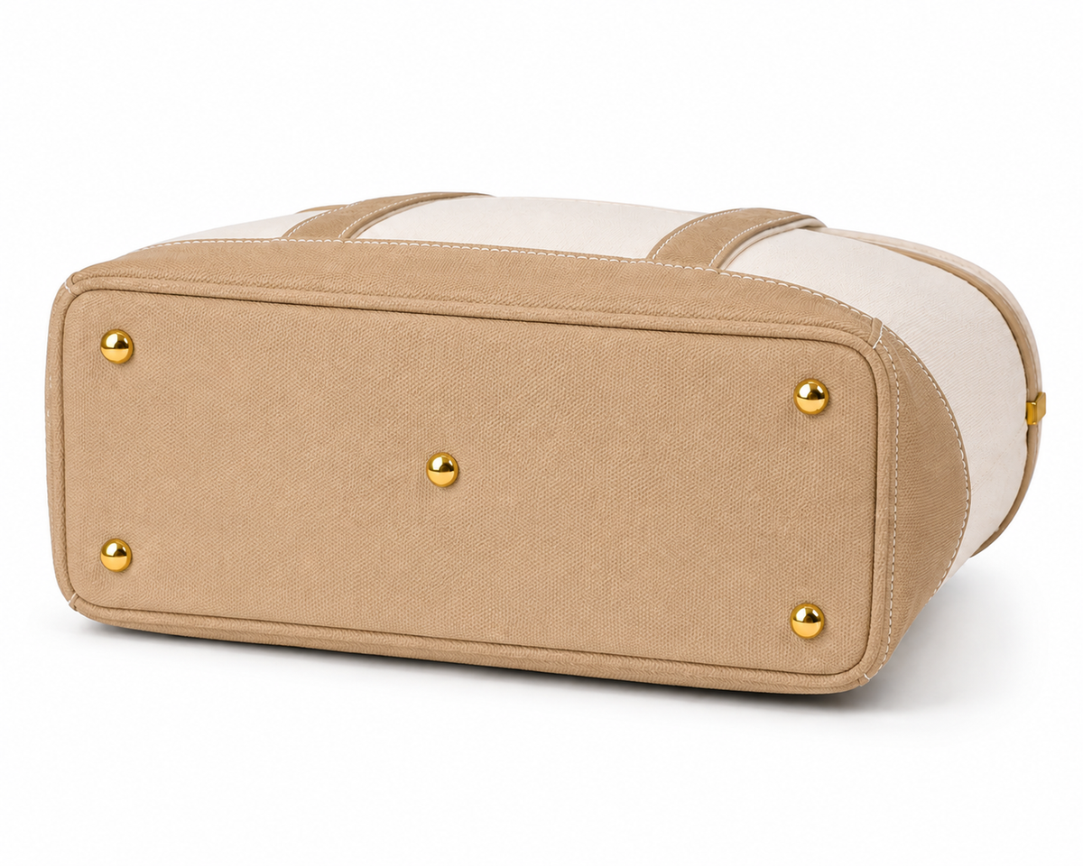 A beige canvas tote bag with cream-colored top panels and gold-tone metal studs, shown from a three-quarters perspective on a white background.