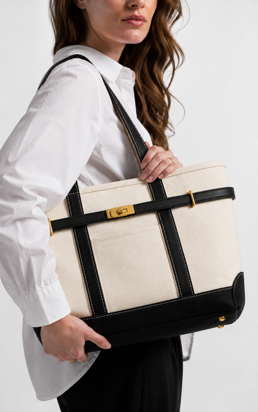 A woman in a white shirt and black pants carries a large cream canvas tote bag with black leather straps and trim, featuring a gold-tone clasp.