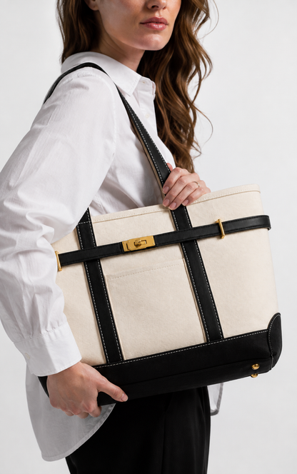 A woman in a white shirt and black pants carries a large cream canvas tote bag with black leather straps and trim, featuring a gold-tone clasp.