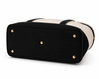 A black and cream canvas tote bag shown from a rear three-quarter angle, featuring a black canvas bottom panel with gold-tone metal studs and a cream canvas upper body with black trim.