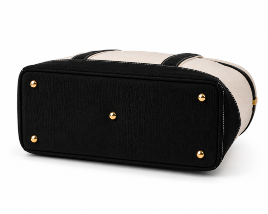 A black and cream canvas tote bag shown from a rear three-quarter angle, featuring a black canvas bottom panel with gold-tone metal studs and a cream canvas upper body with black trim.