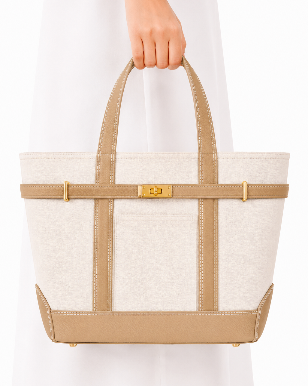 A structured tote bag with an off-white canvas body, beige leather trim and handles, and gold-tone hardware, held by a person wearing a white dress against a plain white background.
