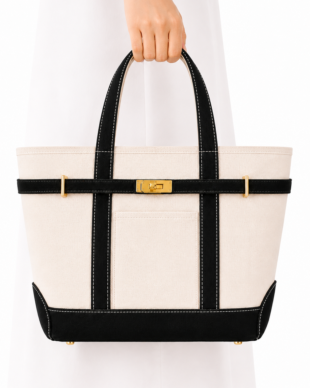 A structured tote bag with an off-white canvas body, black trim, and gold-tone hardware, held by a person wearing a white dress against a plain white background.
