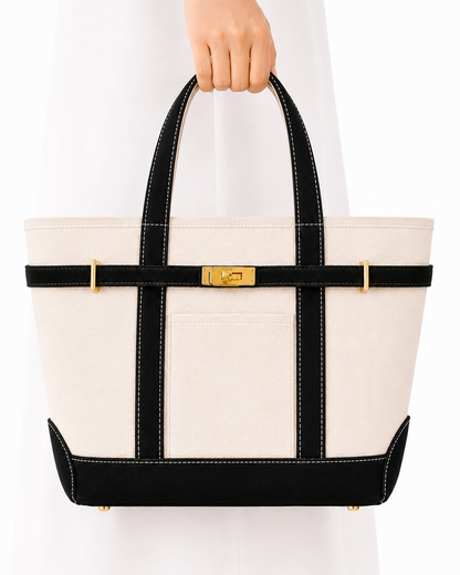 A structured tote bag with an off-white canvas body, black trim, and gold-tone hardware, held by a person wearing a white dress against a plain white background.