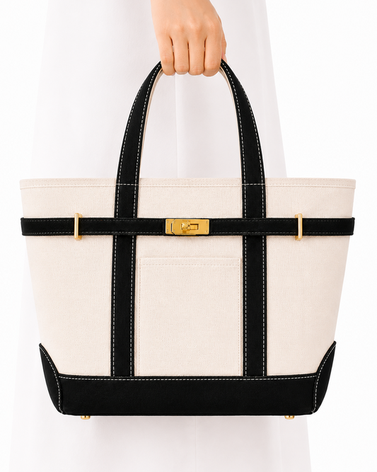 A structured tote bag with an off-white canvas body, black trim, and gold-tone hardware, held by a person wearing a white dress against a plain white background.