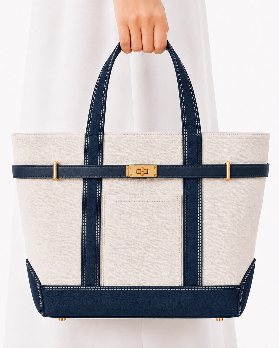 A structured tote bag with an off-white canvas body, navy blue straps and trim, and gold-tone hardware, held by a person wearing a white dress against a clean white background.