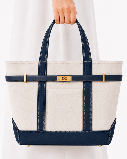 A structured tote bag with an off-white canvas body, navy blue straps and trim, and gold-tone hardware, held by a person wearing a white dress against a clean white background.