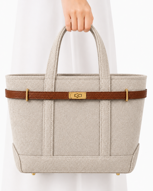 A structured tote bag in light beige felt with a brown leather strap and gold-tone hardware, held by a person wearing a white dress against a clean white background.