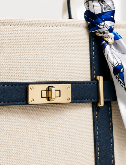 Close-up of a structured tote bag with a cream canvas body, navy blue leather trim, a gold turn-lock clasp, and a blue-and-white patterned scarf tied around the strap.