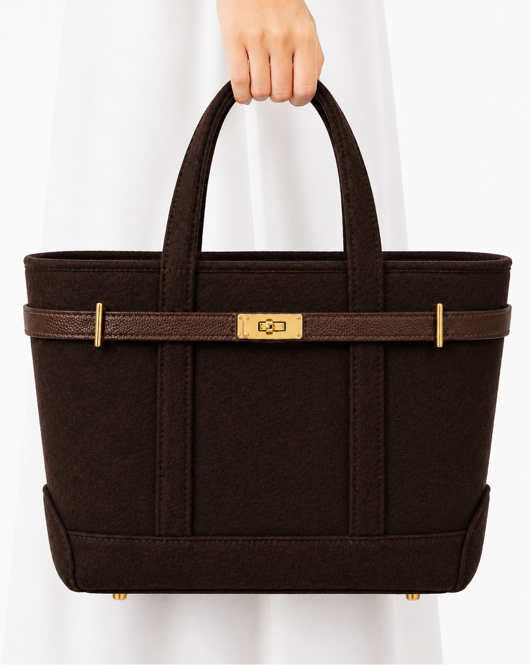 A dark brown felt tote bag with a brown leather strap and gold-tone hardware, held by a person wearing a white dress against a clean white background.